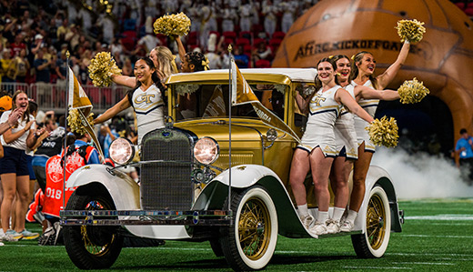 The Ramblin’ Reck, Georgia Tech’s vehicular mascot, enters the football field ahead of a game. Poised on the sides of the Reck are a few of Tech’s cheerleaders.