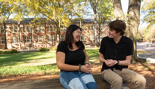 Two Georgia Tech undergraduate students have a conversation on a bench near Harrison Square, a historic part of Tech's Atlanta campus. In the background, a large brick building and foliage are visible.