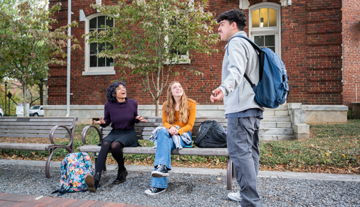Undergraduate students talk on campus in Harrison Square. Two students sit on a bench in front of a brick building, while another student stands nearby.