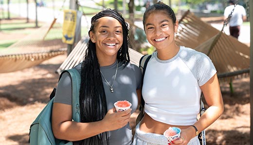 Two undergraduate students at Georgia Tech pose for a photo under a tree near Tech Green on campus. They’re wearing their backpacks and holding snow cones.