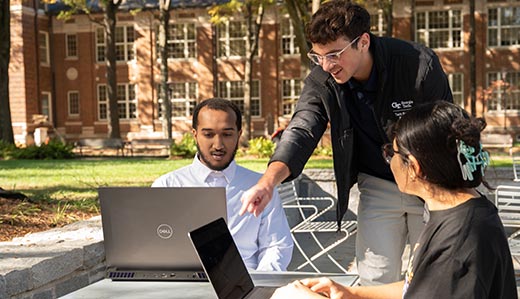 In an outdoor setting, two Georgia Tech students sit at a table with their laptops open while a third student stands and leans forward to point at one of their laptop screens. In the background is a large brick building and several trees.