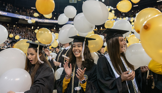 Undergraduate students at Georgia Tech’s graduation ceremony clap and celebrate as white and gold balloons fall from the ceiling of McCamish Pavilion.