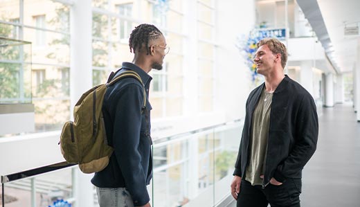 Two Scheller College of Business students are talking in the foreground. The background is the interior of the business school building, which consists of a wall of windows, a blue and yellow glass sculpture suspended from the ceiling, and white accents throughout.