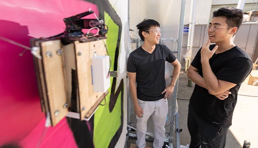 In the background, two College of Computing undergraduate students at Georgia Tech stand and discuss their computing project. In the foreground, their project, housed in a wooden box with wires extending from within, is visible. They appear in an outdoor setting on Tech’s campus.