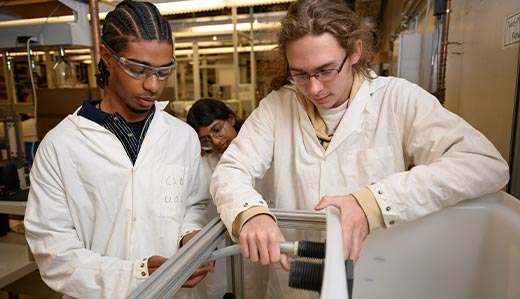 A group of Georgia Tech engineering students wearing protective lab gear, including coats and eyewear, surround a machine. Two students appear to work together to adjust a pipe connected to the machine.