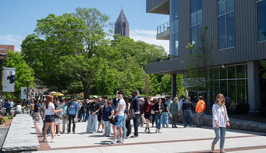 A large gathering of Georgia Tech community members milling around the walkway just outside of the John Lewis Student Center on Tech’s campus in Atlanta. In the background, Atlanta’s iconic ‘pencil building’, nicknamed for its shape and sharp point, is visible. Between the foreground of students and the pencil building is a large swath of tall, full trees. At the edge of the photo, the student center is visible.