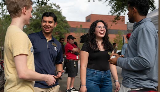 A group of four people have a conversation. One is wearing a polo with the interlocking “GT” logo of Georgia Tech. They appear to be outside on Tech’s campus in Atlanta.