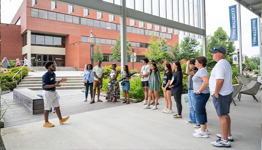 A Georgia Tech undergraduate student gives a tour to a small crowd of visitors on Tech’s campus. The group listens to the tour guide as they examine their surroundings, which consist of a brick building. They are standing under a tall metal awning. Trees and other foliage are visible in the background.