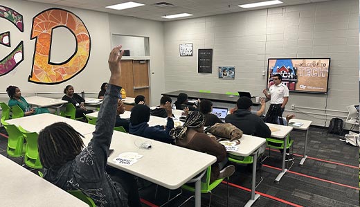 In a classroom setting, a student near the camera raises their hand to ask a question. At the front of the room, a Georgia Tech representative is standing next to a projected presentation with Tech Tower on it.