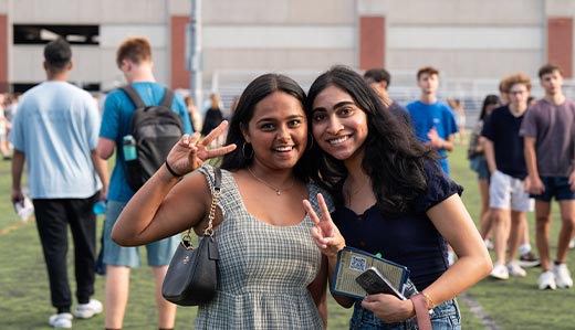 A close-up photo of two Georgia Tech undergraduate students posing for a photo on campus, smiling and holding up peace signs. Blurred in the background is a larger group of Georgia Tech students. Some are walking, some are standing, and some have backpacks on.
