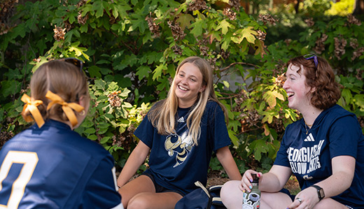 Three students sit on a picnic blanket in a green space beneath Tech Tower on campus. They are talking and laughing.