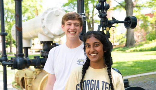 Two students pose with the old gold, black, and white steam engine on Georgia Tech’s campus. Trees and other greenery are in the background.