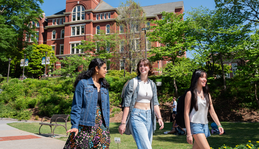 In the foreground, three Georgia Tech undergraduate students talk as they walk on the green beneath Tech Tower on Georgia Tech’s Atlanta campus. Behind them, the front of the base of Tech Tower is visible, as well as lush greenery and foliage in front of the building.